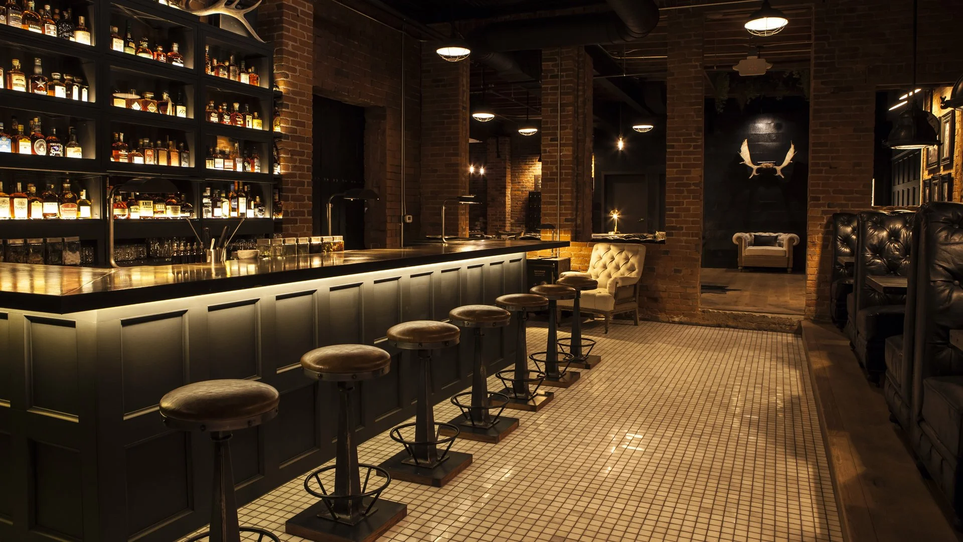Dimly lit bar with a long black counter, high stools with round leather seats, brick walls, shelves of liquor bottles behind the bar, and cozy seating areas with plush chairs and a mirror reflecting the space.