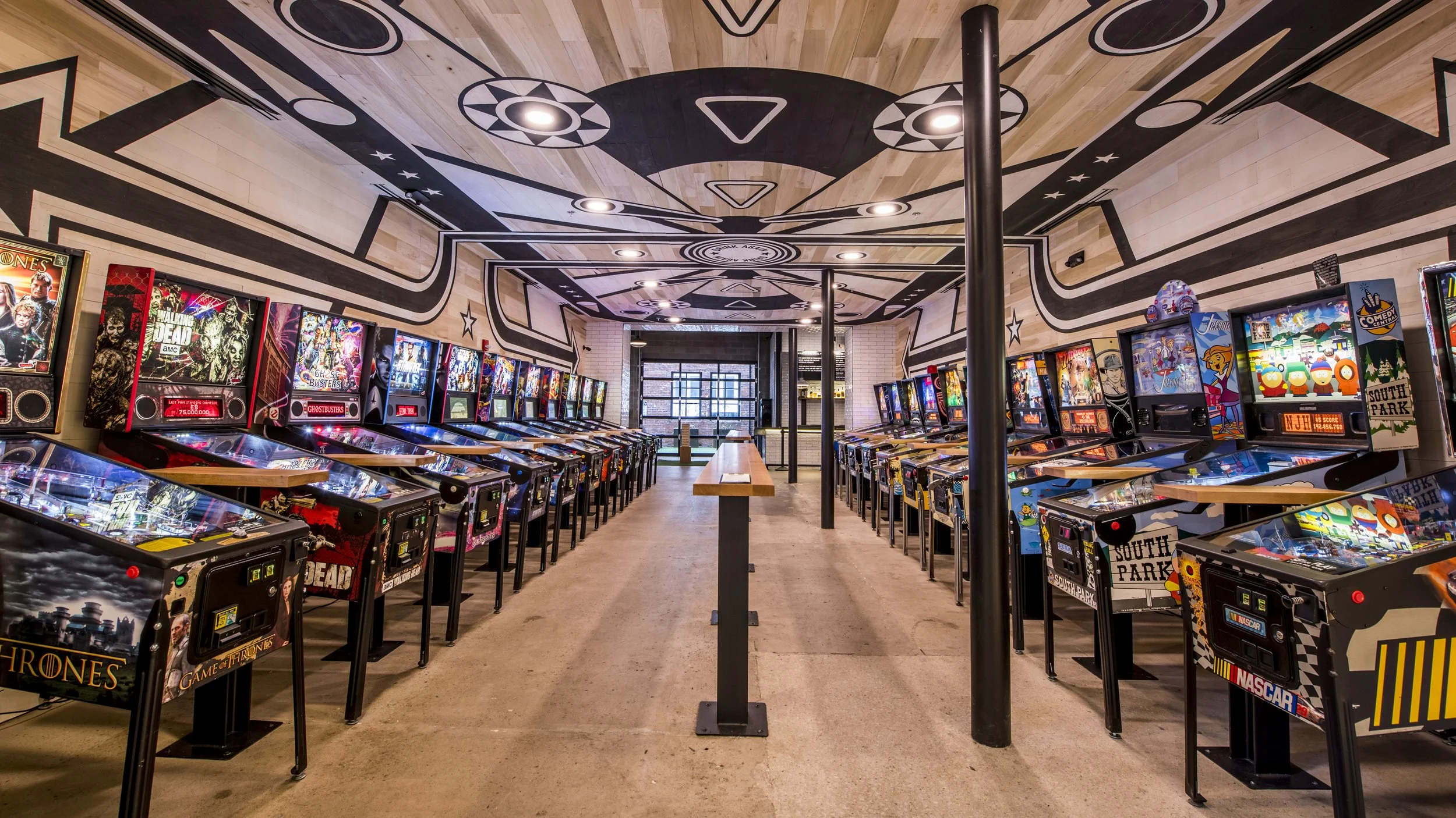 Interior of a pinball arcade with two rows of classic pinball machines, themed after popular shows and characters, with a central walkway and decorative ceiling.