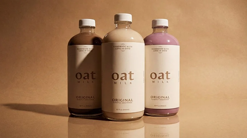 Three bottles of oat milk, one chocolate, one original, and one berry flavor, on a reflective surface with a beige background.