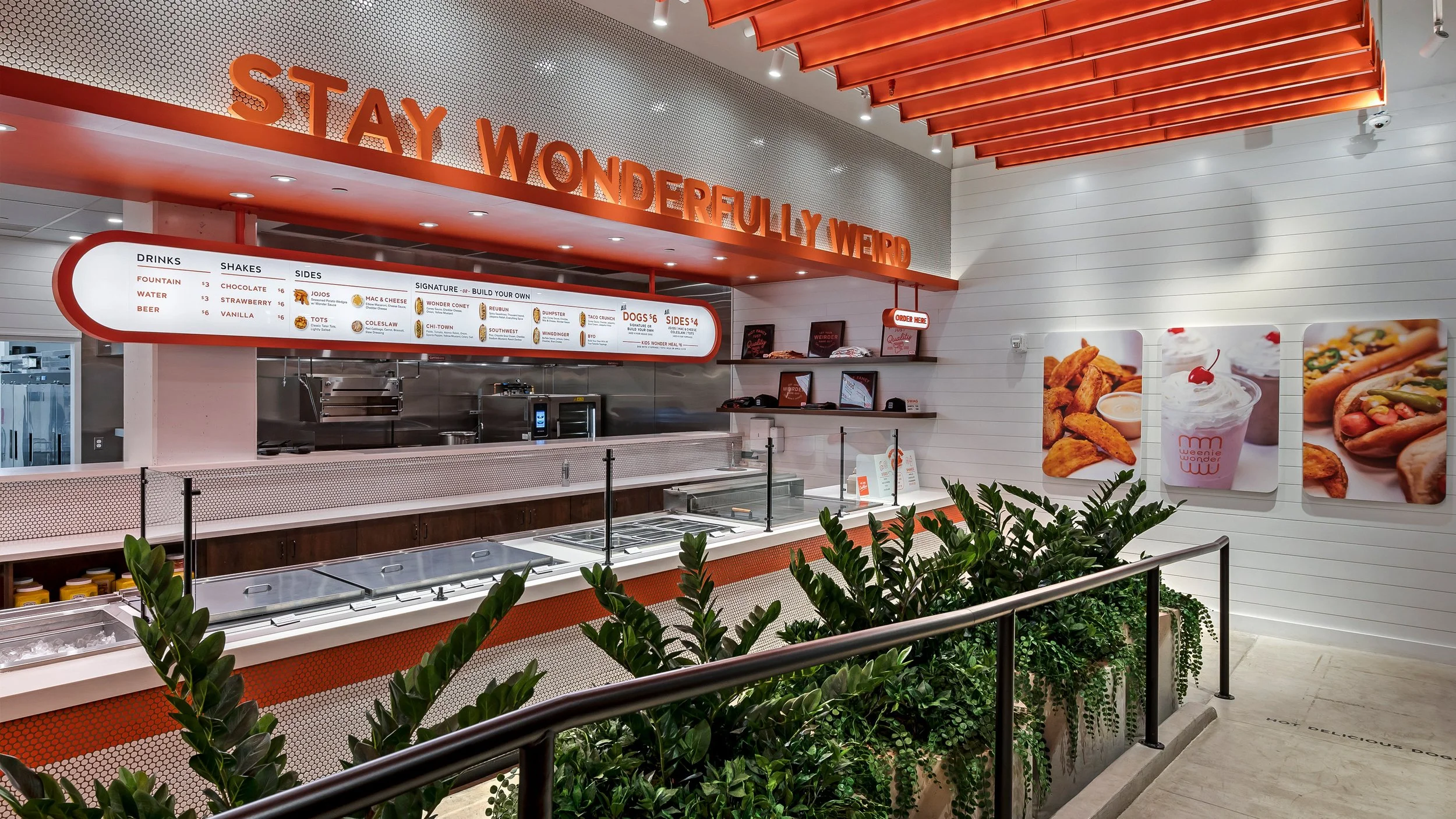Inside a fast-food restaurant with a modern design, menu and promotional images on the wall, and a row of plants along the front counter.