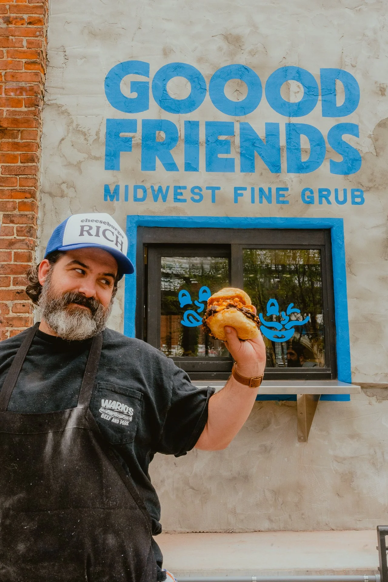 A man with a beard and mustache wearing a blue and white cap and black shirt holding a sandwich inside a restaurant with a sign that reads 'Good Friends Midwest Fine Grub' in blue letters.