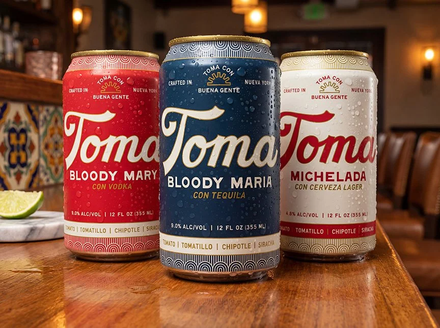 Three cans of Toma craft beer on a wooden table with lime wedge and restaurant interior in background