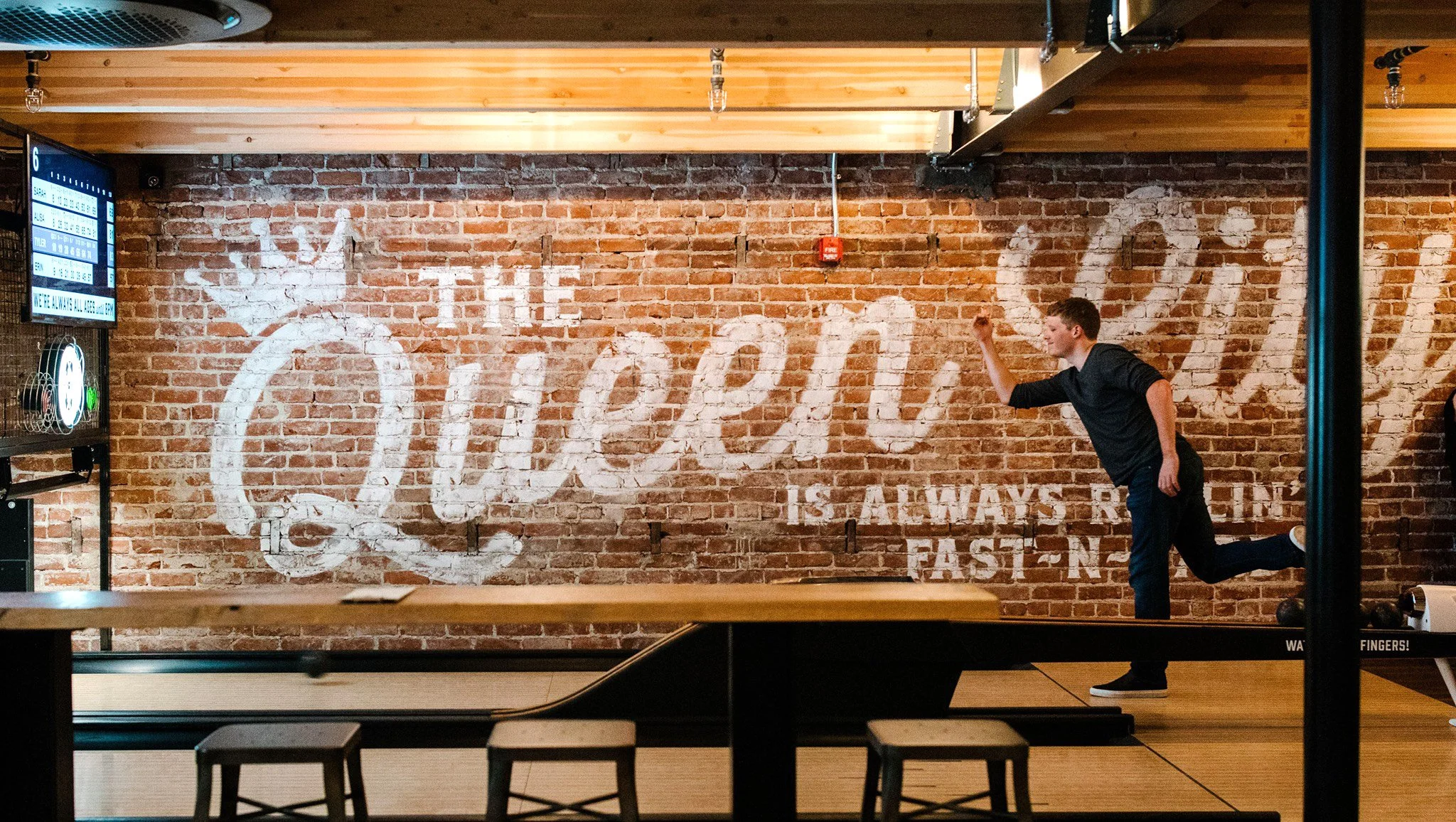 A young man playing ping pong in an indoor recreational space with a brick wall in the background and a large white painted Coca-Cola logo.