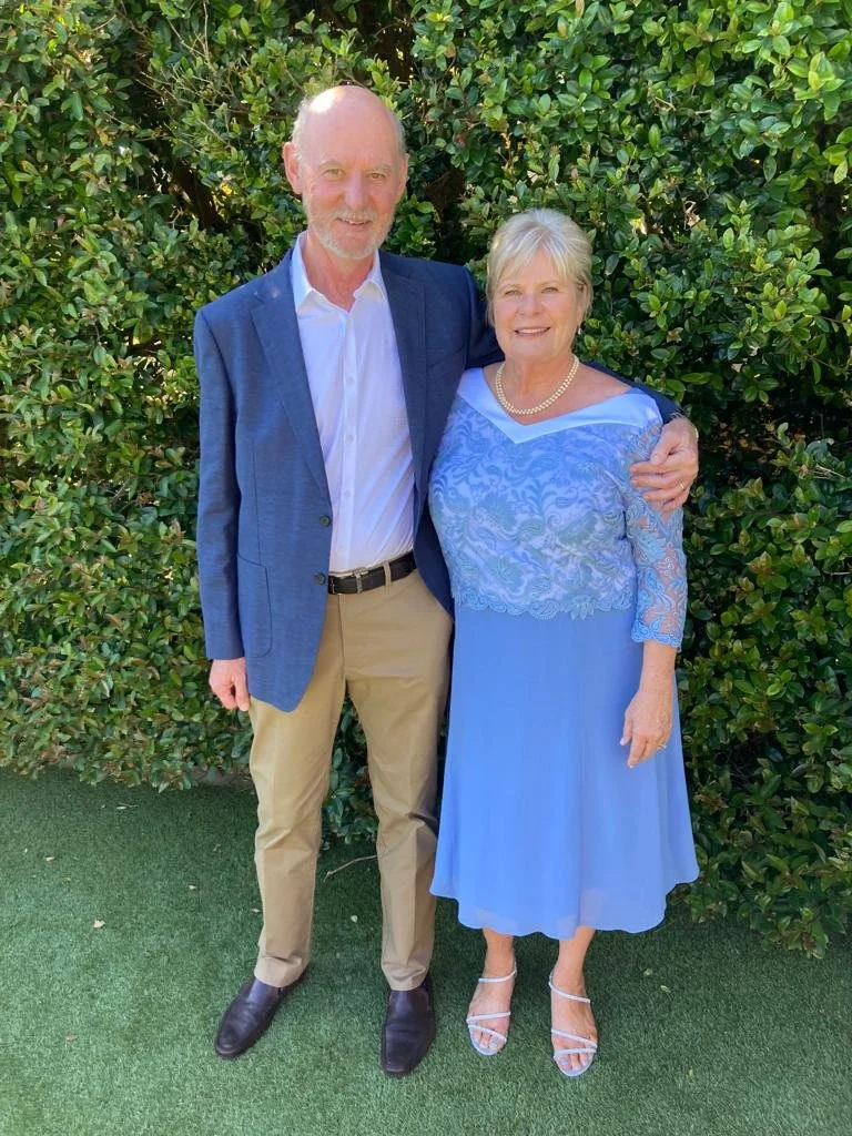 A man and woman standing outdoors in front of green shrubbery. The man is wearing a blue blazer, white shirt, khaki pants, and black shoes. The woman is wearing a blue dress with lace details, pearl necklace, and white strappy heels. They are smiling