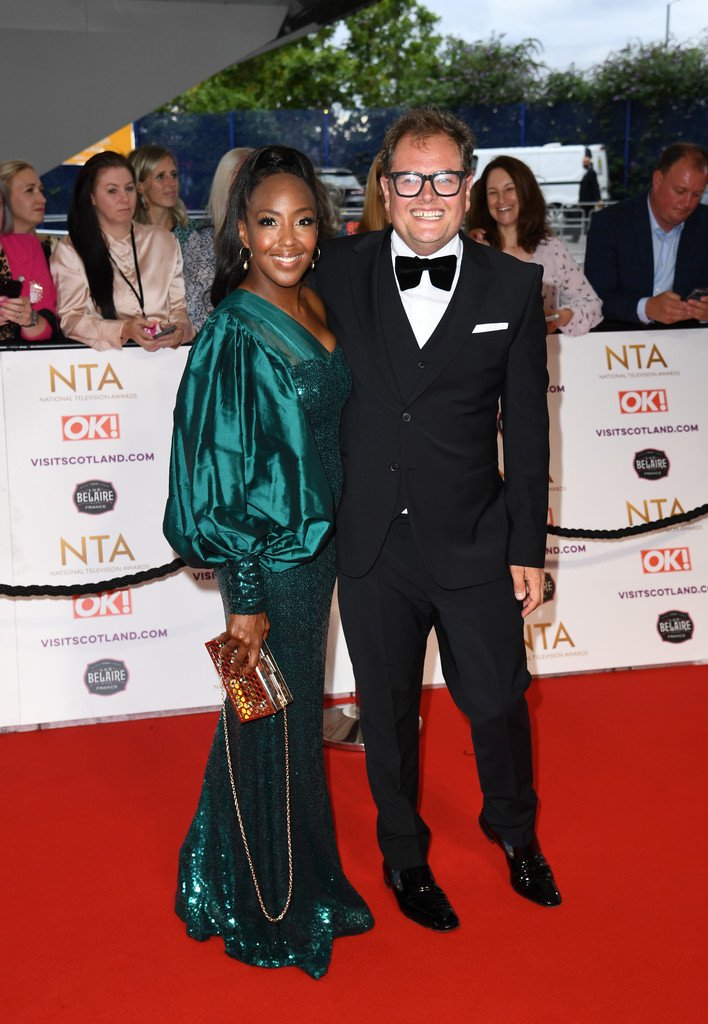 Two people, a woman in a teal sequin gown and a man in a black tuxedo, standing on a red carpet at an event with other guests and banners in the background.