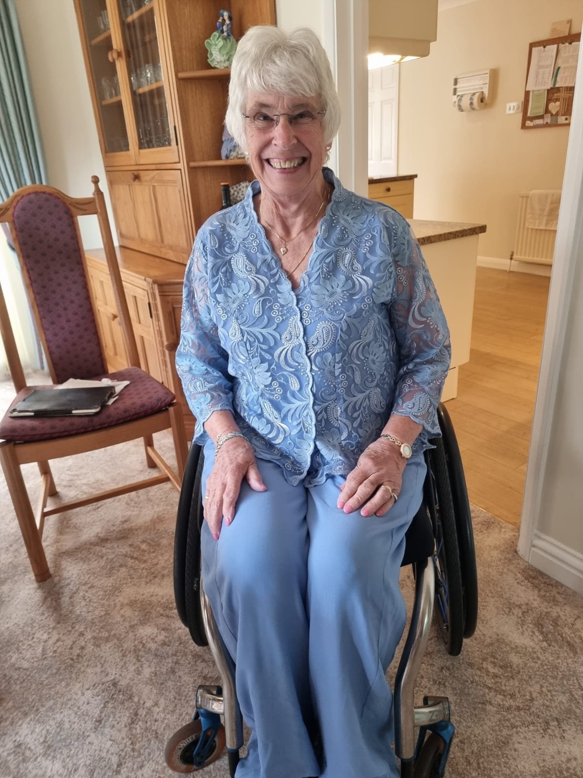 An elderly woman with gray hair smiling and sitting in a wheelchair indoors. She is wearing a light blue patterned blouse and matching pants, with jewelry including a bracelet, watch, and necklace. Background shows wooden furniture, a chair with a bo