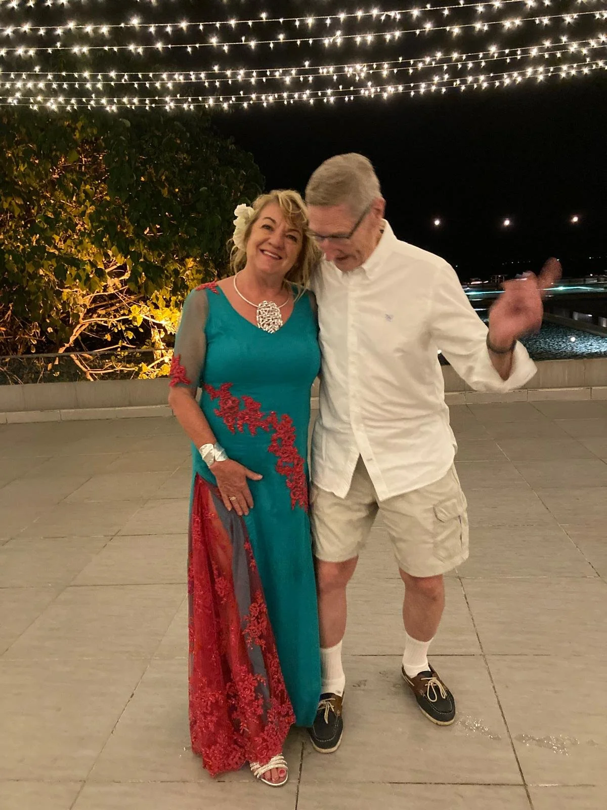 A woman in a teal dress with red embroidery and a man in a white shirt and shorts at night under string lights, smiling and dancing together.
