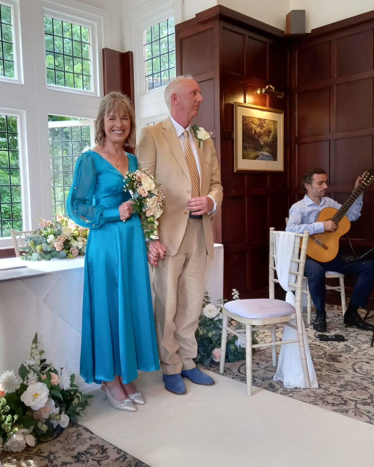 A smiling woman in a bright blue dress holding a bouquet of flowers standing next to a man in a light beige suit with a flower pinned to his lapel, holding hands during a wedding ceremony. A guitarist playing guitar is seated nearby, and floral arran