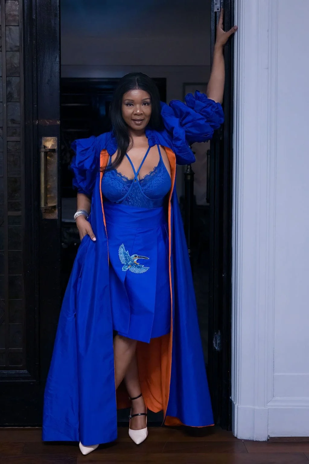 A woman standing in a doorway wearing a vibrant blue dress with a bird embroidered on the skirt, layered with a matching jacket and lace top, accessorized with white high heels and silver bangles, posing confidently.