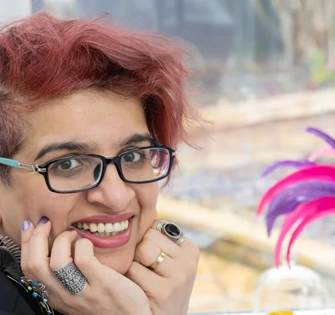 A woman with short, pinkish hair, glasses, and a bright smile, resting her face on her hands near colorful feathers in the background.