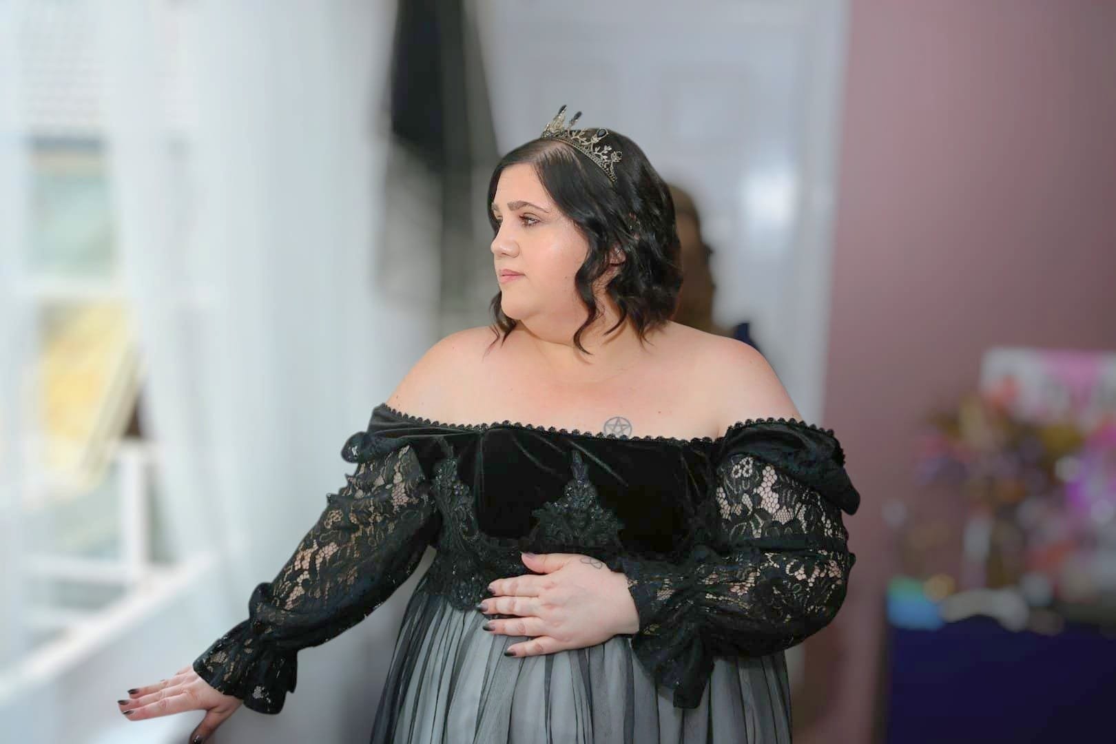 A woman dressed in a black off-shoulder lace gown with a tiara standing indoors, facing to the left with her hand on a wall.