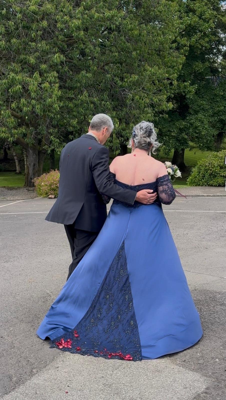 A couple dressed in formal attire, the man in a black suit and the woman in a blue gown with lace details, standing outdoors on a paved area with trees and bushes in the background.