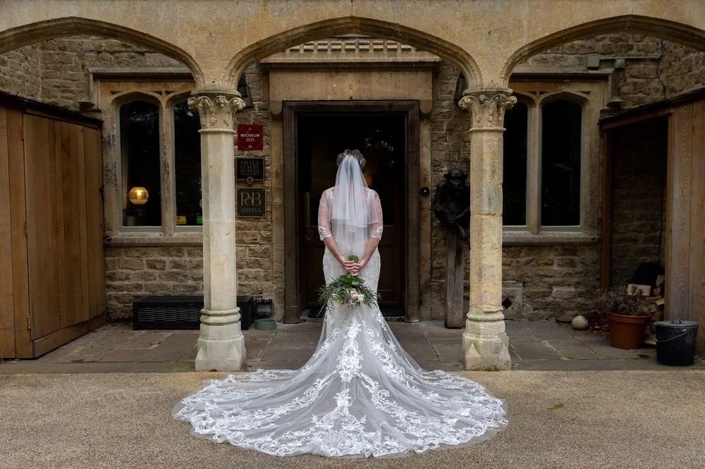 Bride in a white wedding dress with a long train and veil, standing with her back to the camera in front of a stone building with arches, holding a bouquet of flowers behind her back.