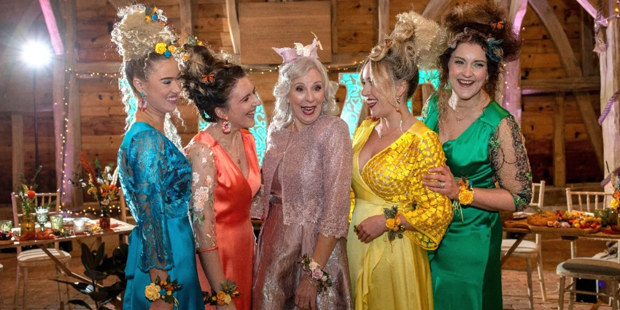 Group of six women dressed in colorful, fancy dresses at a festive indoor celebration, smiling and talking. The setting appears to be a decorated rustic barn with tables, chairs, and floral arrangements.