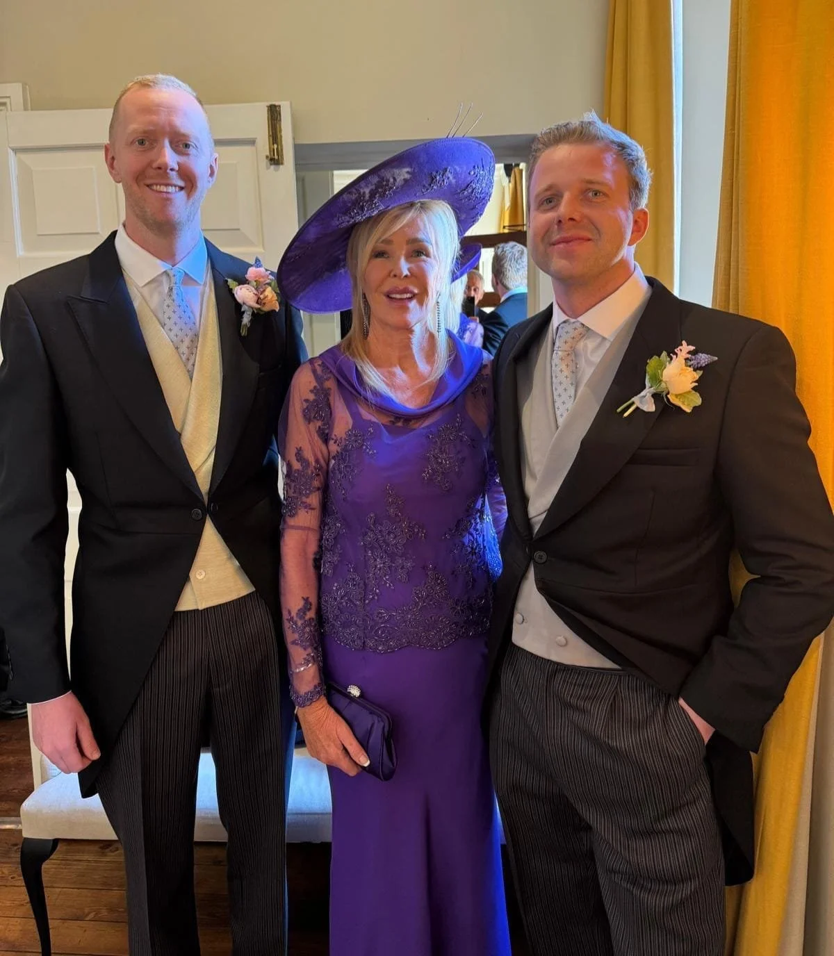 Three people dressed in formal attire at a wedding or similar event, two men in tuxedos with boutonnières and a woman in a purple dress and hat, standing indoors with yellow curtains.