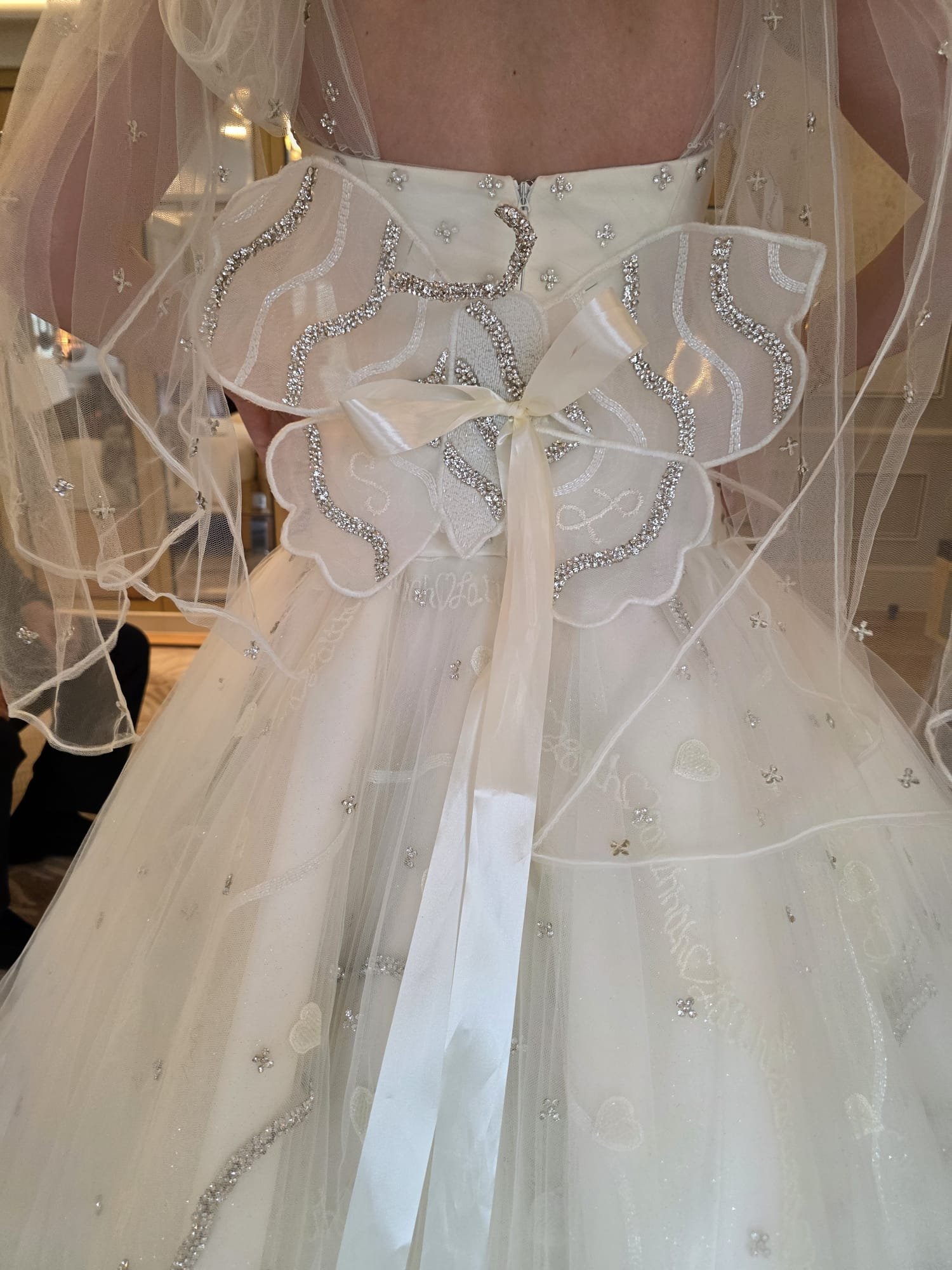 Close-up of a white wedding dress with rhinestone and pearl embellishments, a satin bow, and an overlay of sheer fabric with small flower and star details.