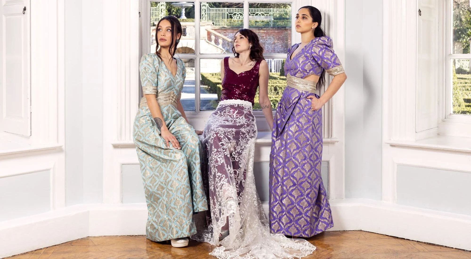 Three women wearing elegant dresses are posed indoors near a large window with a garden view. One woman in a teal and gold dress is seated on the window sill, another in a purple and gold dress stands to her right with hand on hip, and the third in a