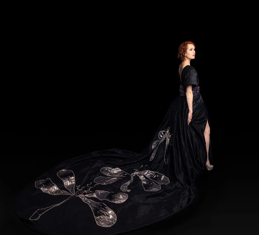 A woman with red hair, dressed in a black gown with a long train decorated with silver insects, standing against a black background.