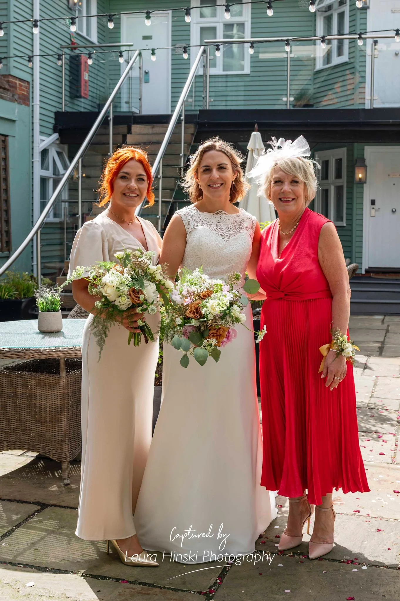Three women at a wedding, dressed in elegant gowns, holding floral bouquets, standing outdoors with string lights overhead, in front of a green house with steps and a glass railing.