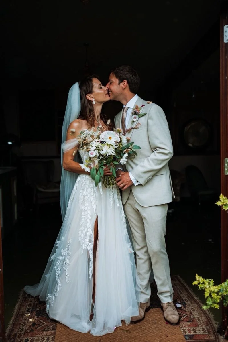 A bride and groom sharing a kiss during their wedding ceremony, with the bride holding a bouquet of flowers and wearing a lace wedding dress and a veil, while the groom is dressed in a light-colored suit.