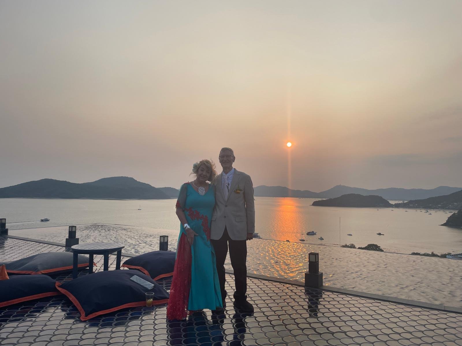 A couple dressed in formal attire standing on a tiled patio near a body of water at sunset. The woman wears a turquoise and red gown with floral details, and the man wears a light beige suit. They are smiling with the setting sun behind them, casting