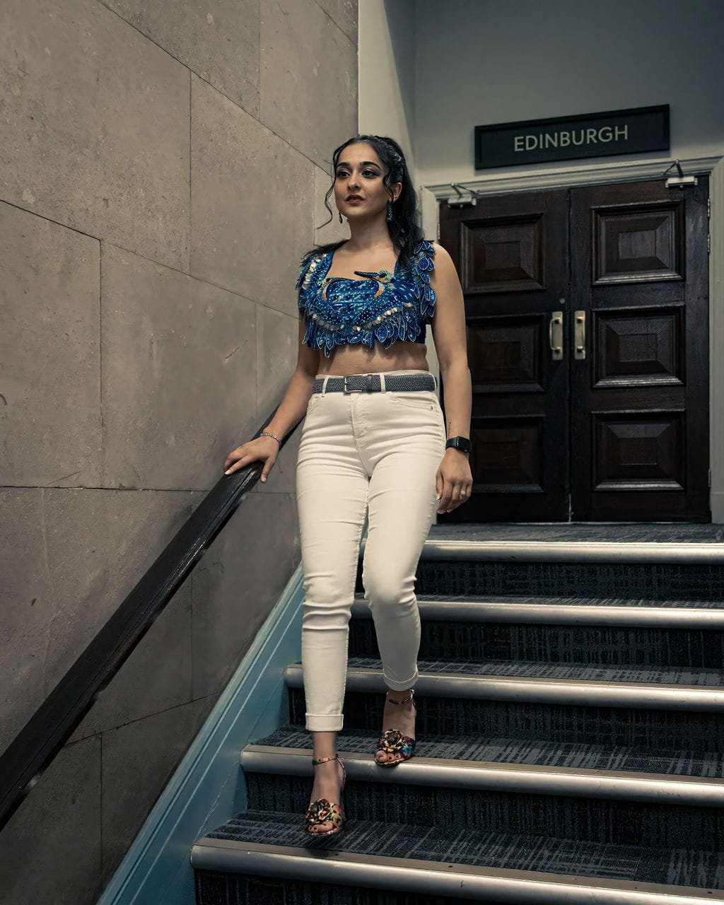 A woman in a blue embroidered crop top and white pants stands on indoor staircase near a wall and a door with an Edinburgh sign above it.