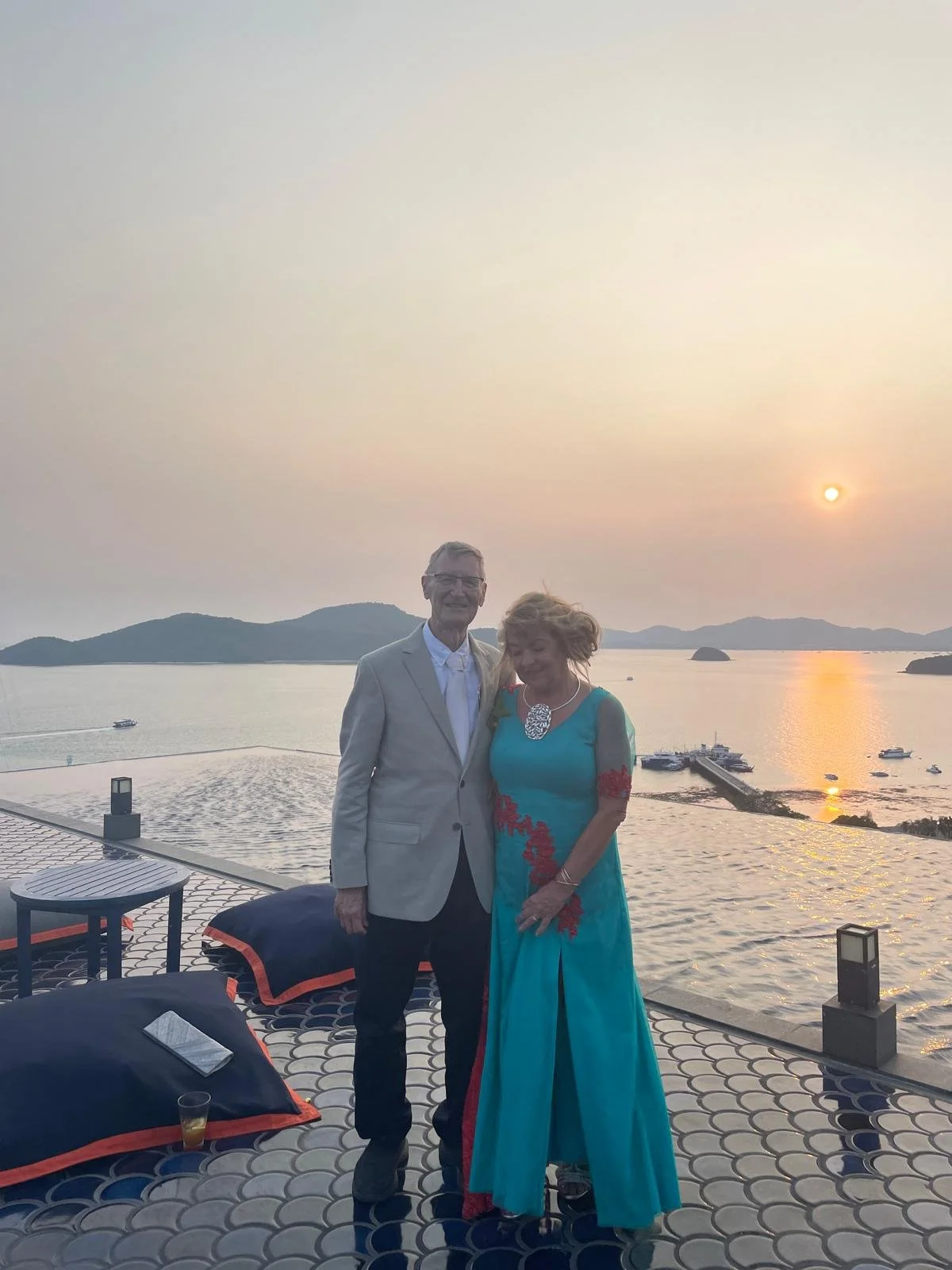 A man and woman standing on a terrace overlooking a body of water with boats, mountains in the background, during sunset. The man wears a light-colored suit, and the woman wears a turquoise dress with red floral details and a large necklace.