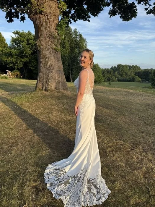 A woman in a white wedding dress standing outdoors near a large tree, smiling and looking at the camera with a scenic background of grass, trees, and a blue sky.
