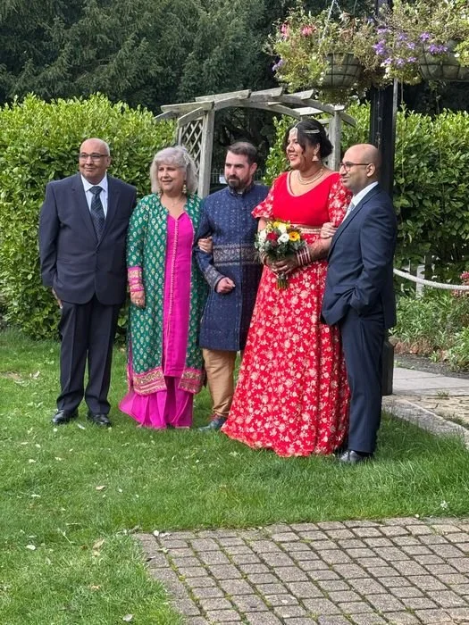 Group of five people at a wedding, standing outdoors on grass near a wooden arch with flowers, with trees and bushes in the background. The bride is in a red dress holding a bouquet, and the groom and others are dressed in formal attire.