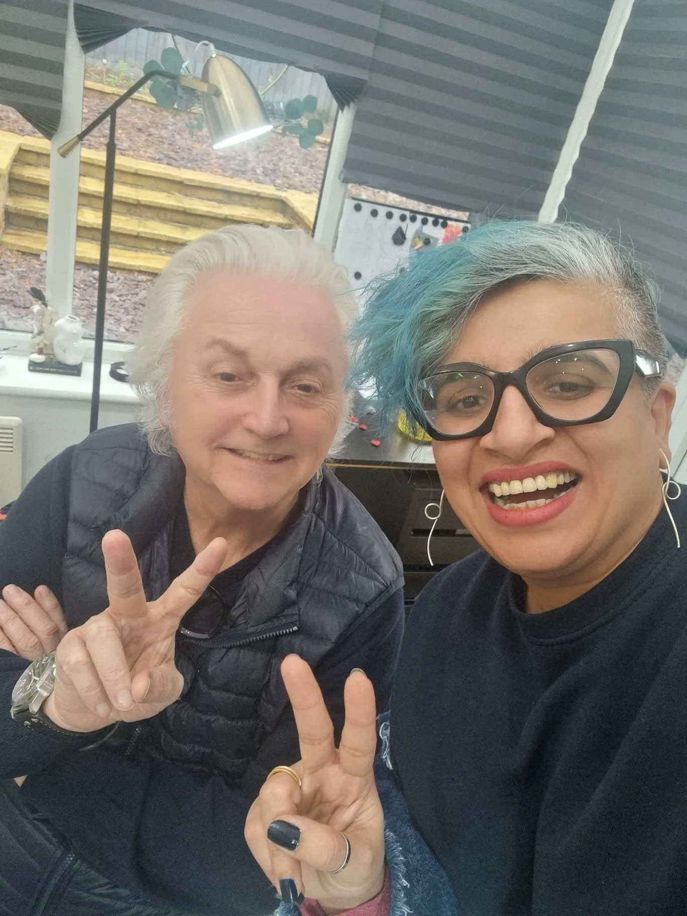 Two people smiling and making peace signs, one with white hair and the other with teal-colored hair and black glasses, inside a room with a window and some plants outside.