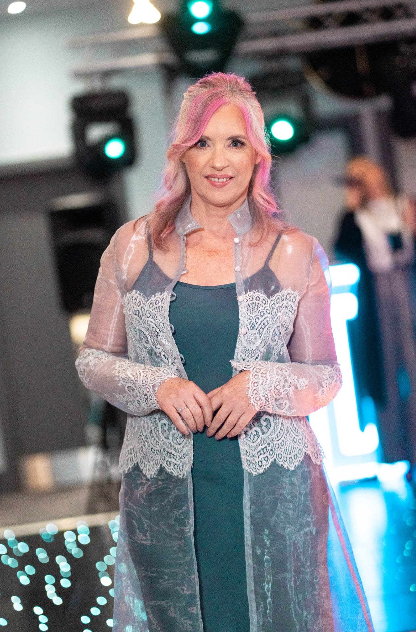 A woman with pink hair styled in loose waves, wearing a teal top and a lace-embellished sheer jacket, standing in front of a colorful light background at an indoor event.