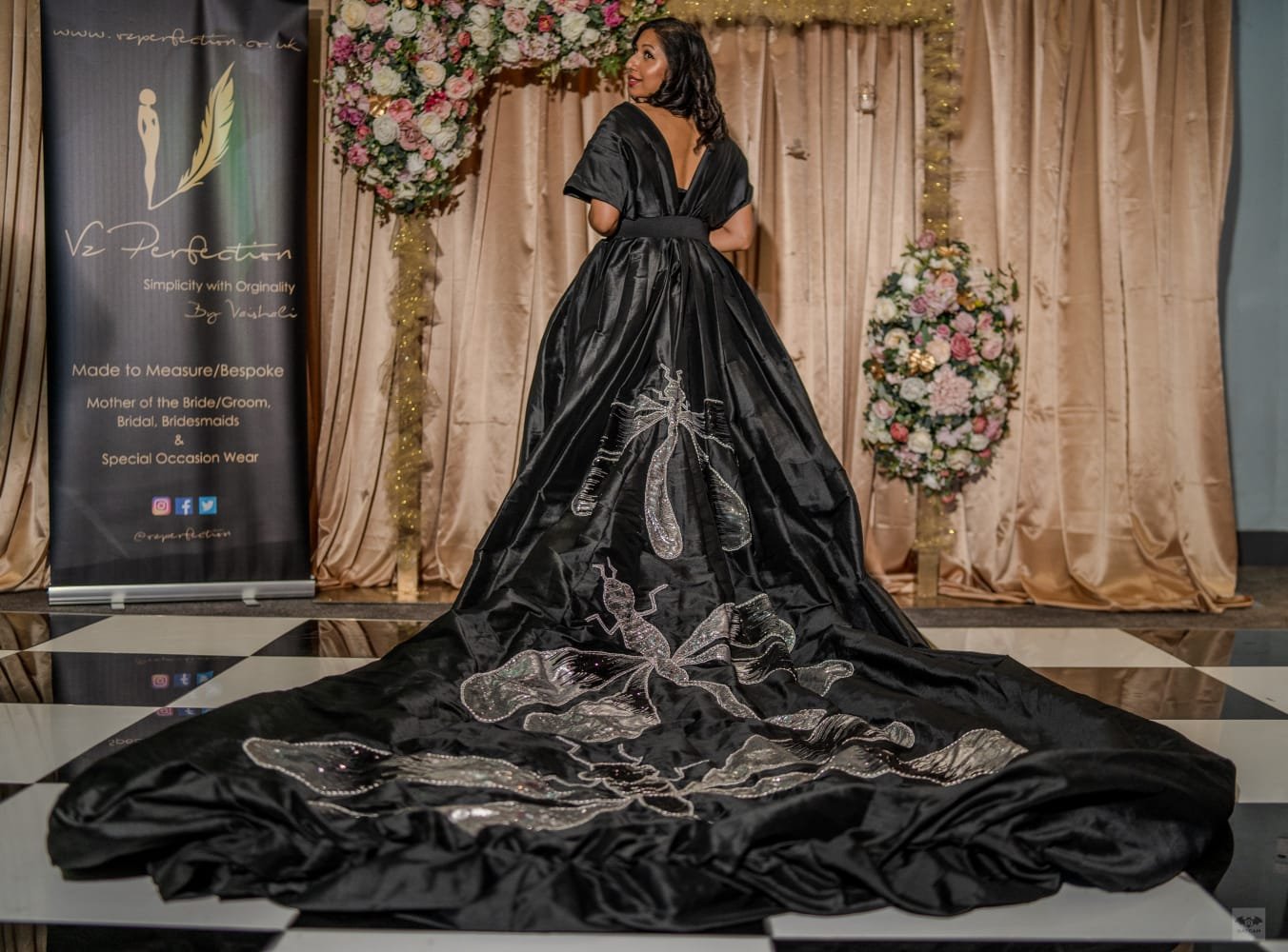 A woman in a black gown with silver embroidery stands on a checkered floor in front of a pink curtain and floral arrangements, showcasing the back of her formal dress.