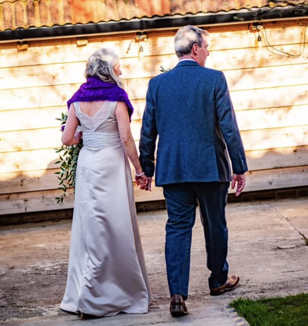 A couple holding hands, walking away from camera, in wedding attire, outside near wooden wall.