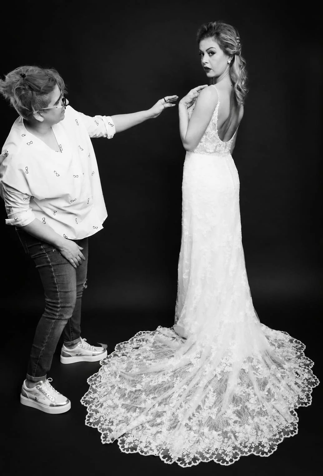 A woman in a wedding dress being assisted or styled, with another person reaching towards her, against a dark background.