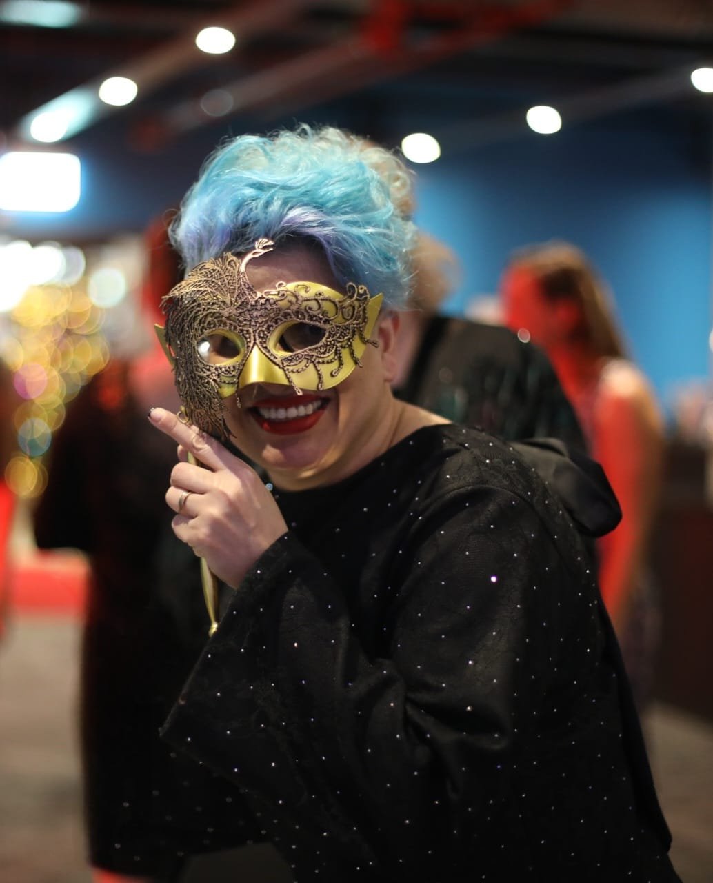 Woman with dyed blue and purple hair wearing a black sparkly jacket and a gold decorative mask, smiling at a festive event.