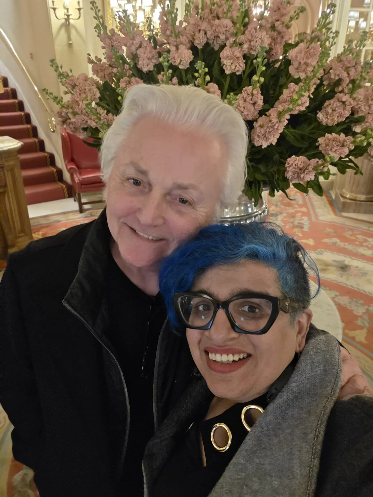 Two people are taking a selfie in an elegant, well-lit room with a large floral arrangement of pink flowers and green foliage in the background. The person on the left has light skin, white hair, and is smiling, while the person on the right has dark