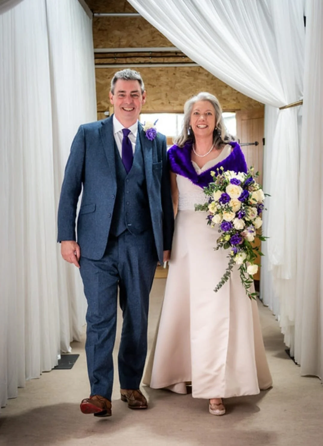 A man and woman in wedding attire walking together through a decorated archway, smiling, with the woman holding a bouquet of white and purple flowers. The setting appears to be indoors with draped curtains and a rustic wooden ceiling.