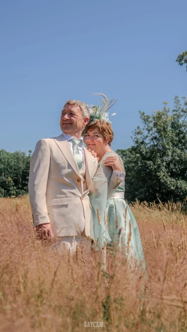 A happy couple dressed in wedding attire standing in a field with tall grass under a clear blue sky. The man wears a light-colored suit with a white shirt and tie, and the woman wears a pastel-colored dress with a fascinator on her head. They are smi