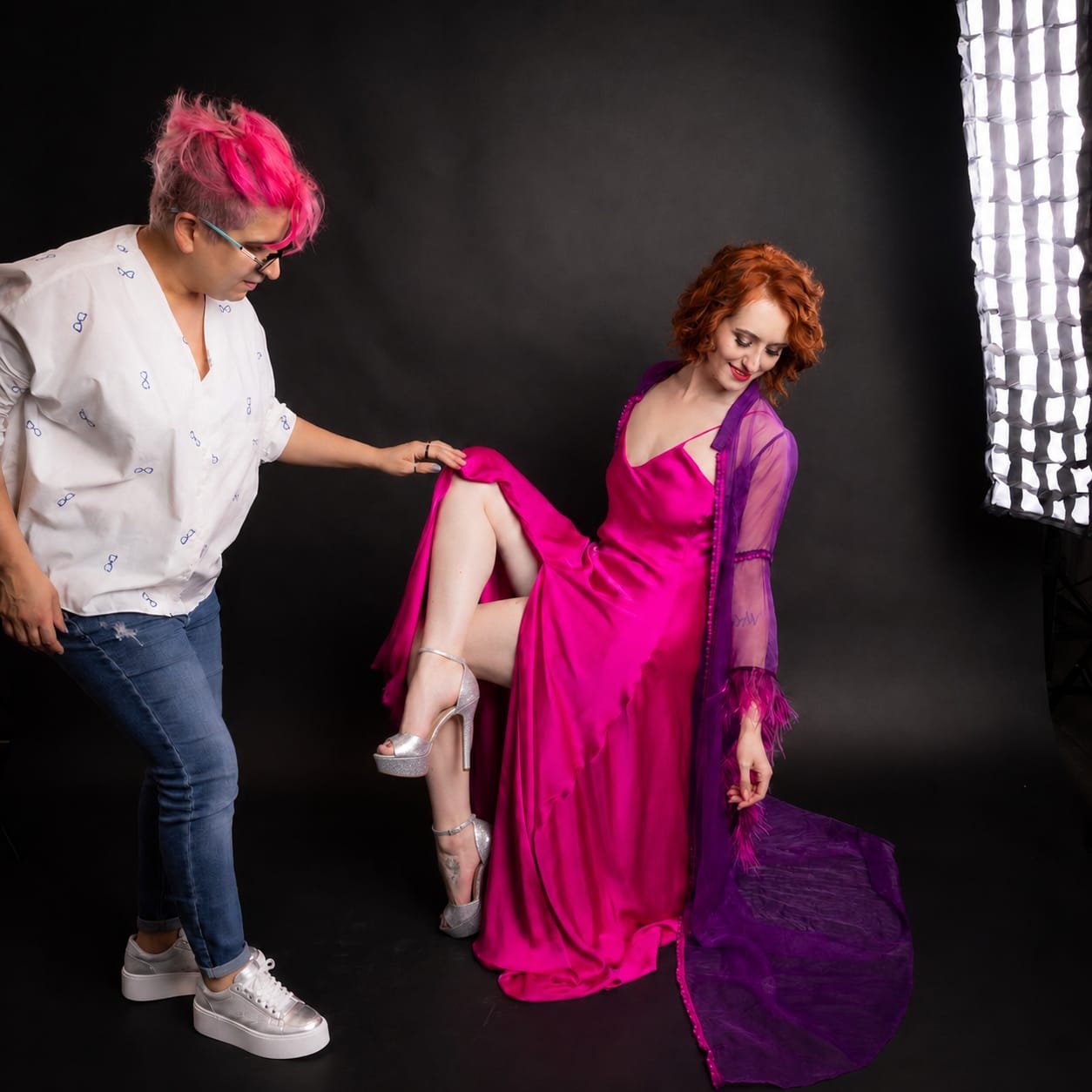 A woman in a vibrant pink dress with purple sheer sleeves and a purple sheer shawl is posing with her leg raised, showing off silver high-heeled shoes. She is sitting against a black background and smiling while a stylist with pink hair and glasses a