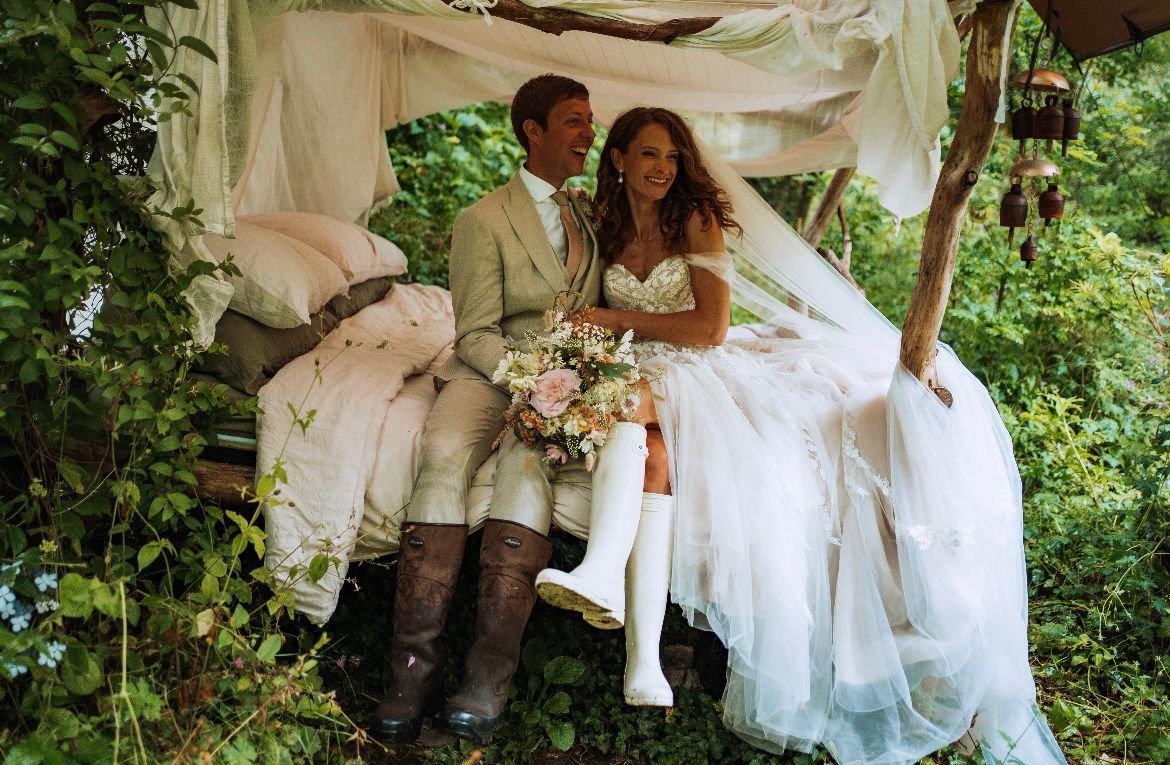 A happy bride and groom sitting together on a rustic bed outdoors, surrounded by greenery. The bride is in a white wedding dress and the groom in a beige suit, holding a bouquet of flowers. They are smiling and appear joyful.