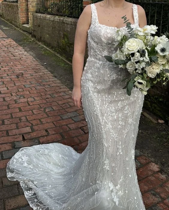 A bride wearing a white wedding dress holding a bouquet of white and green flowers, standing on a brick sidewalk.