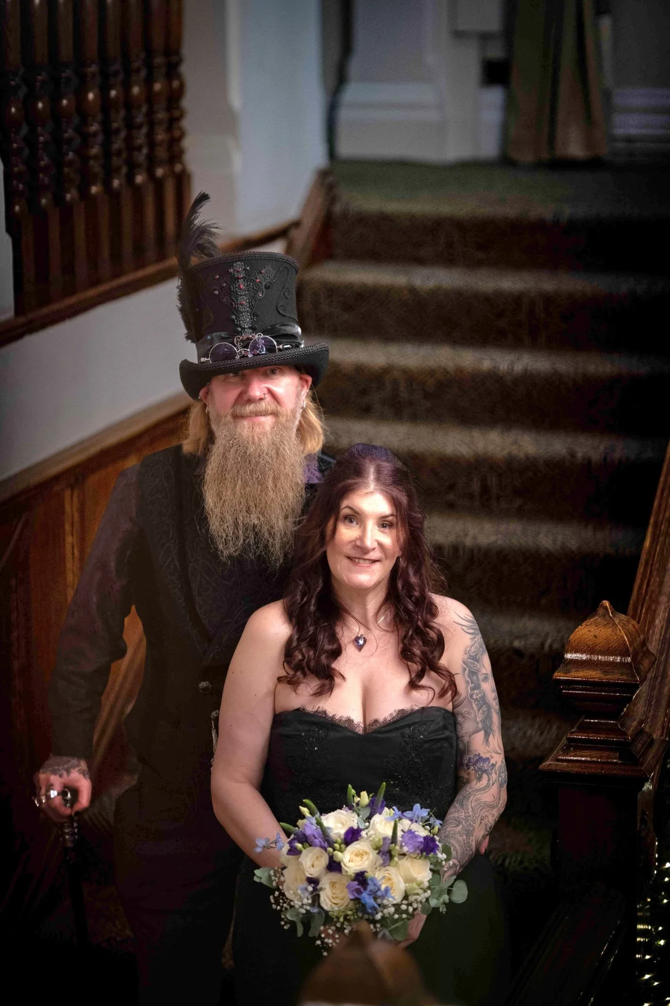 A woman in a strapless black dress holding a bouquet of white and purple flowers, standing in front of a man with long beard wearing a moustache and a black hat with feathers, inside a house with stairs and piano.