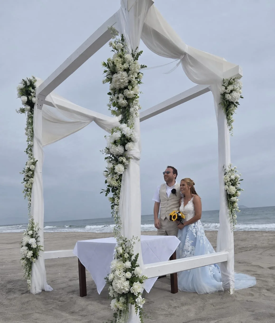 A bride and groom standing under a white wedding arch decorated with white flowers and drapes, on a beach with the ocean in the background.