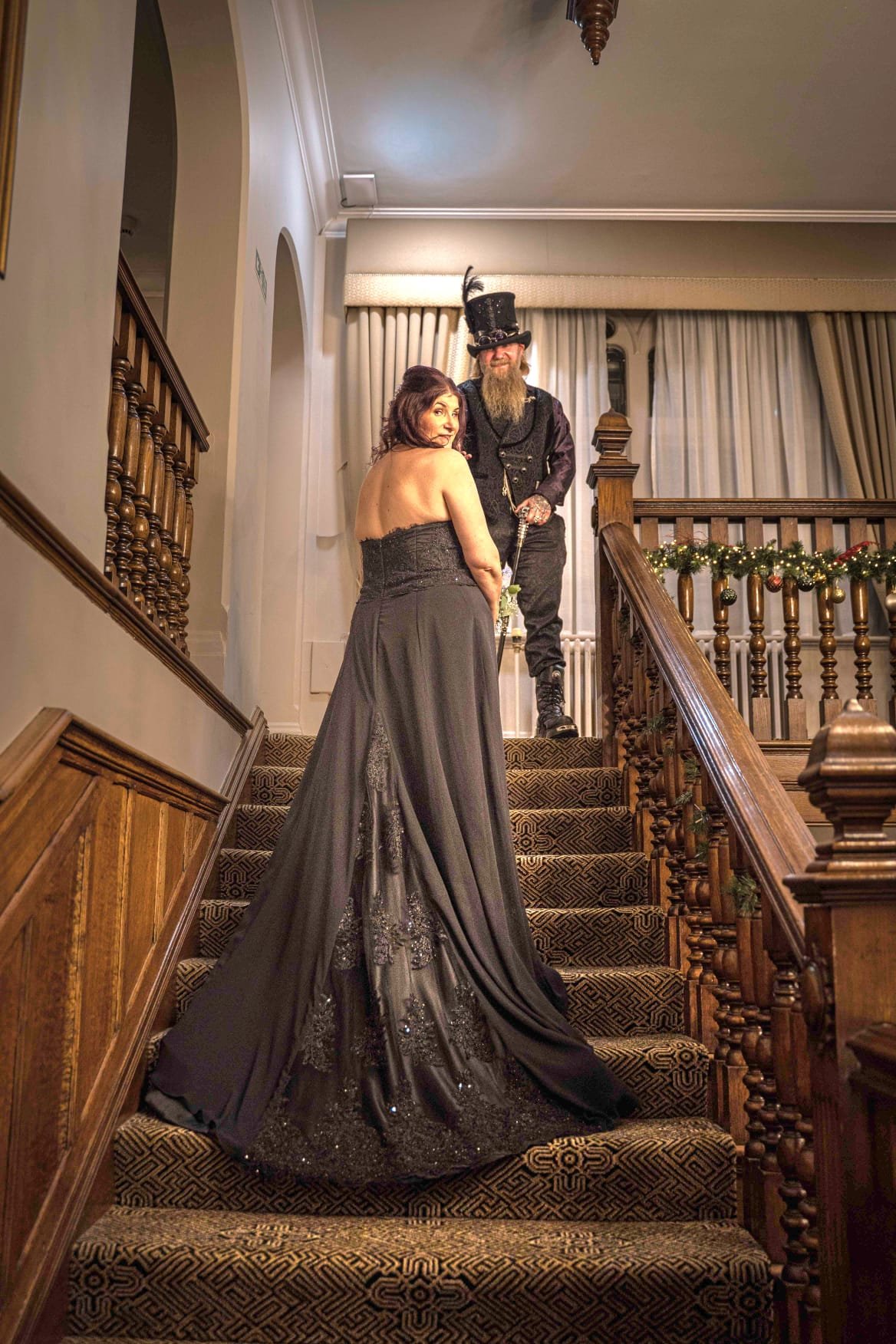A woman in a strapless black gown with detailed embroidery stands on a staircase, with a man in a Victorian-style costume, including a top hat with a feather, standing behind her. The staircase is decorated with green garland, and the setting looks f