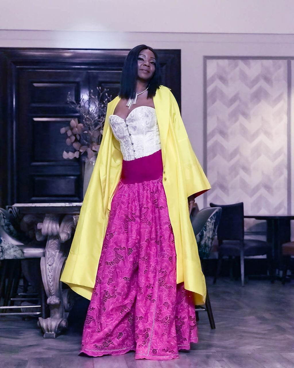A woman in a white corset, bright pink lace pants, and a yellow coat posing indoors.