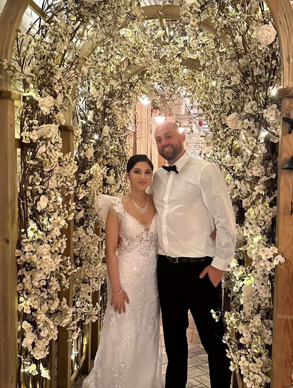 A bride and groom standing under a floral arch at their wedding reception. The bride wears a white lace wedding gown and the groom wears a white shirt with a black bow tie. They are smiling and embracing each other.