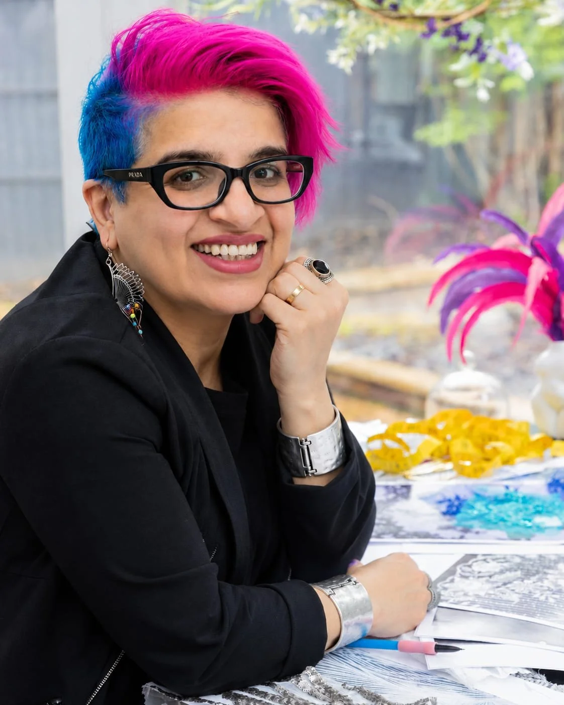 A woman with short, colorful hair (pink and blue) wearing black glasses, a black jacket, and earrings, smiling at the camera while sitting at a table with art supplies, colorful leaves, and plants in the background.