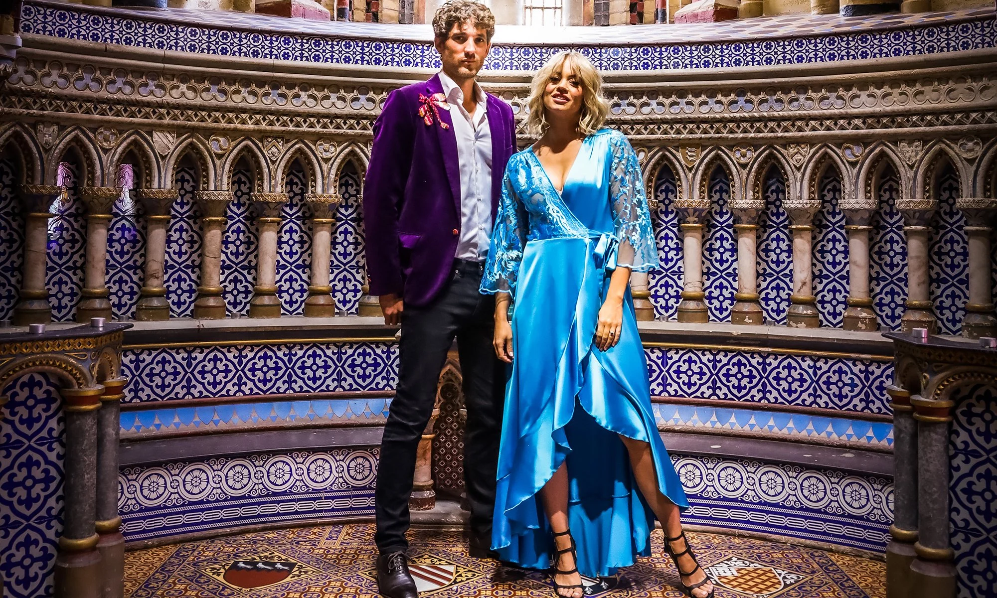 A man and a woman standing together inside a building with ornate blue and gold tiled walls. The man wears a purple velvet blazer with an embroidered detail on his shoulder, a white shirt, and black pants. The woman wears a bright blue satin dress wi