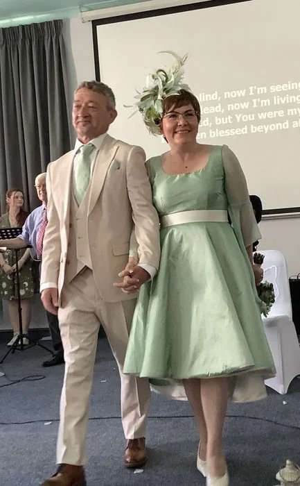 A couple dressed in formal attire holding hands, walking in front of a group inside a decorated room, likely at a wedding or celebration. The woman is wearing a green vintage-style dress with a matching floral headpiece, and the man is in a light-colored suit with a mint green tie.