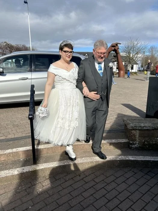 A bride in a white dress and a man in a suit, possibly at a wedding ceremony, walking together outdoors on a paved area with a parked van and other people in the background.
